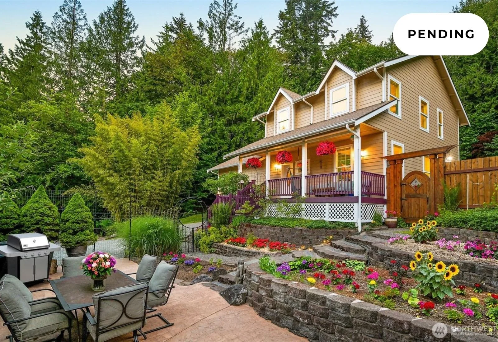 A two-story house with a wraparound porch, surrounded by a lush, green, forested landscape, with a garden and patio area in the foreground featuring colorful flowers and patio furniture.