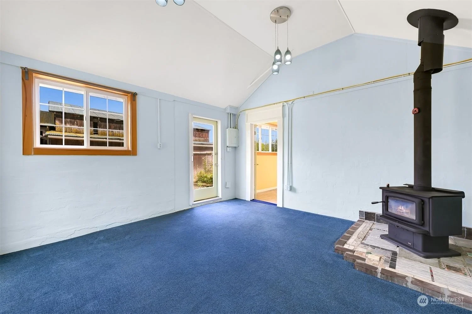 Empty living room with blue carpet, white brick walls, a window with wooden trim, a door leading outside, and a wood stove on a tiled hearth.
