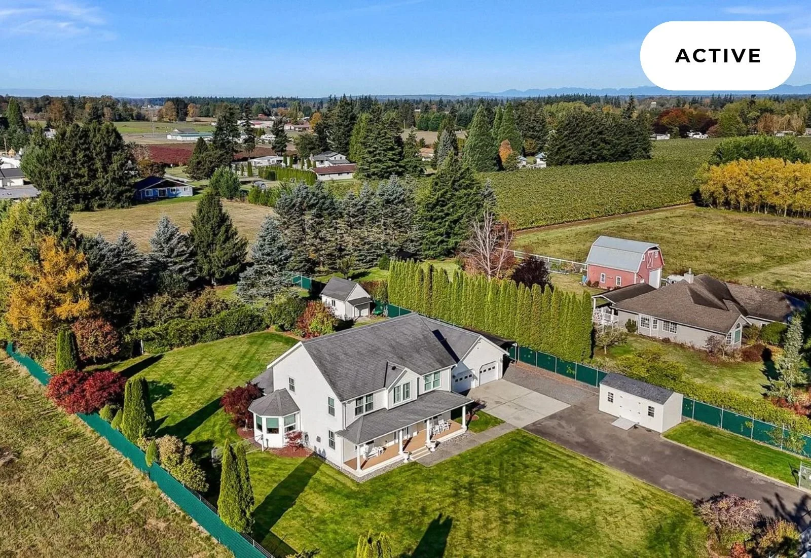 Aerial view of a suburban neighborhood with a white two-story house with a porch, a gray roof, and a green backyard, surrounded by trees and a green fence, with other houses, trees, and fields in the background under a blue sky labeled 'ACTIVE'.