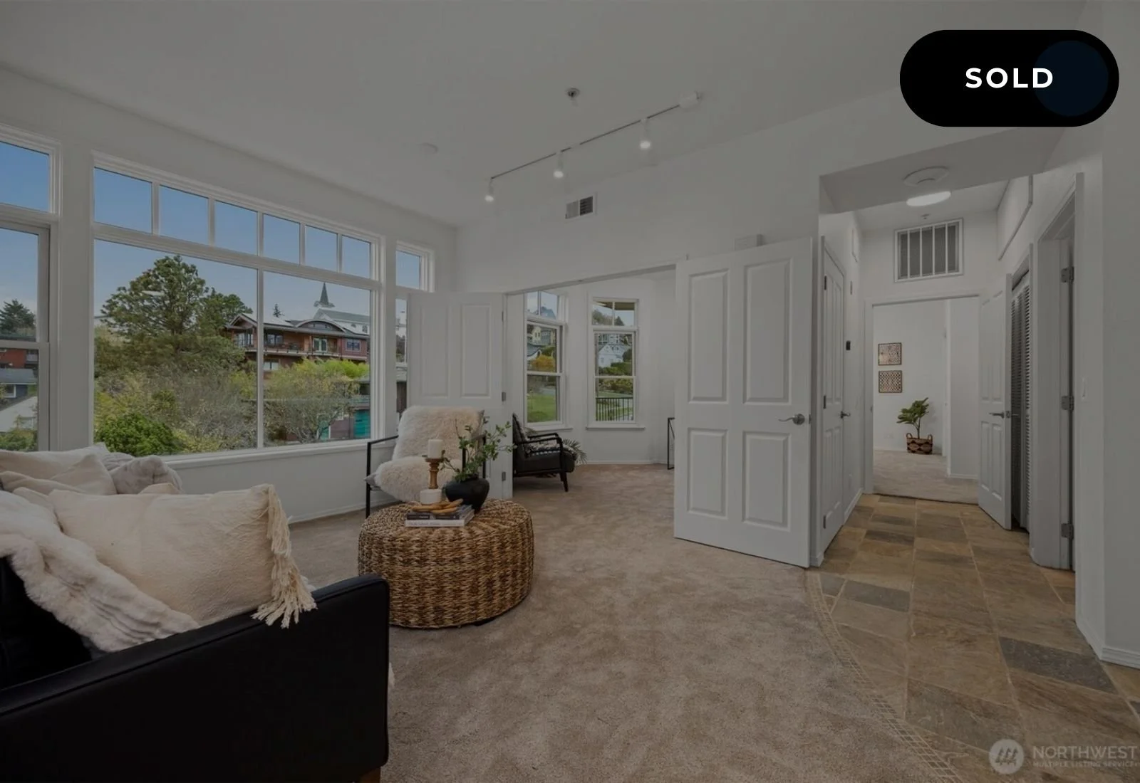 Bright living room with large windows, white walls, beige carpet, black sofa with white throw pillows, round woven coffee table with a black vase and books, and two chairs near the windows.