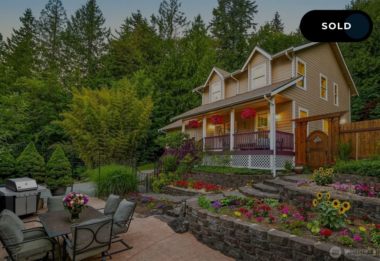 A two-story house with a wraparound porch, surrounded by a lush, green, forested landscape, with a garden and patio area in the foreground featuring colorful flowers and patio furniture.