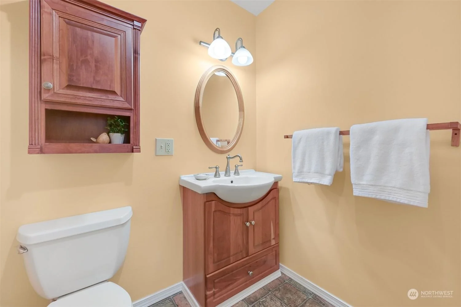 Small bathroom with a wooden cabinet on the wall, a mirror above the sink, white towels on a wooden towel rack, a toilet, and a small cabinet below the sink. The walls are painted yellow.