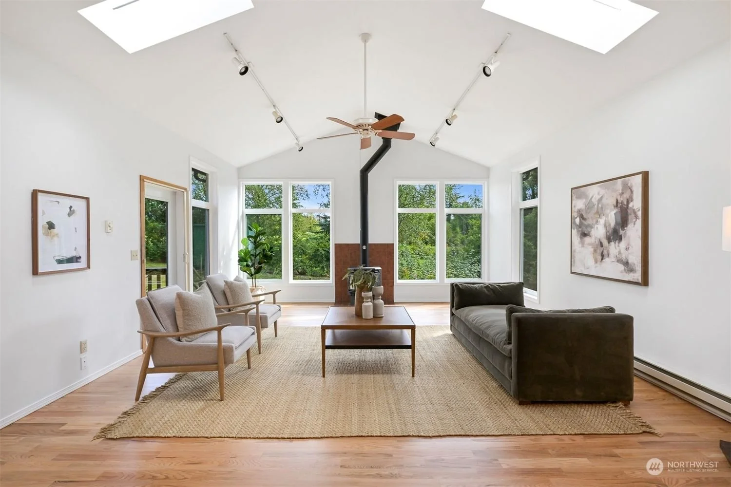 Bright living room with white walls, large windows, wooden floor, and a vaulted ceiling with skylights. Contains a gray sofa, three beige armchairs, a wooden coffee table with white vases, a potted plant, a wood-burning stove with a black pipe, and a