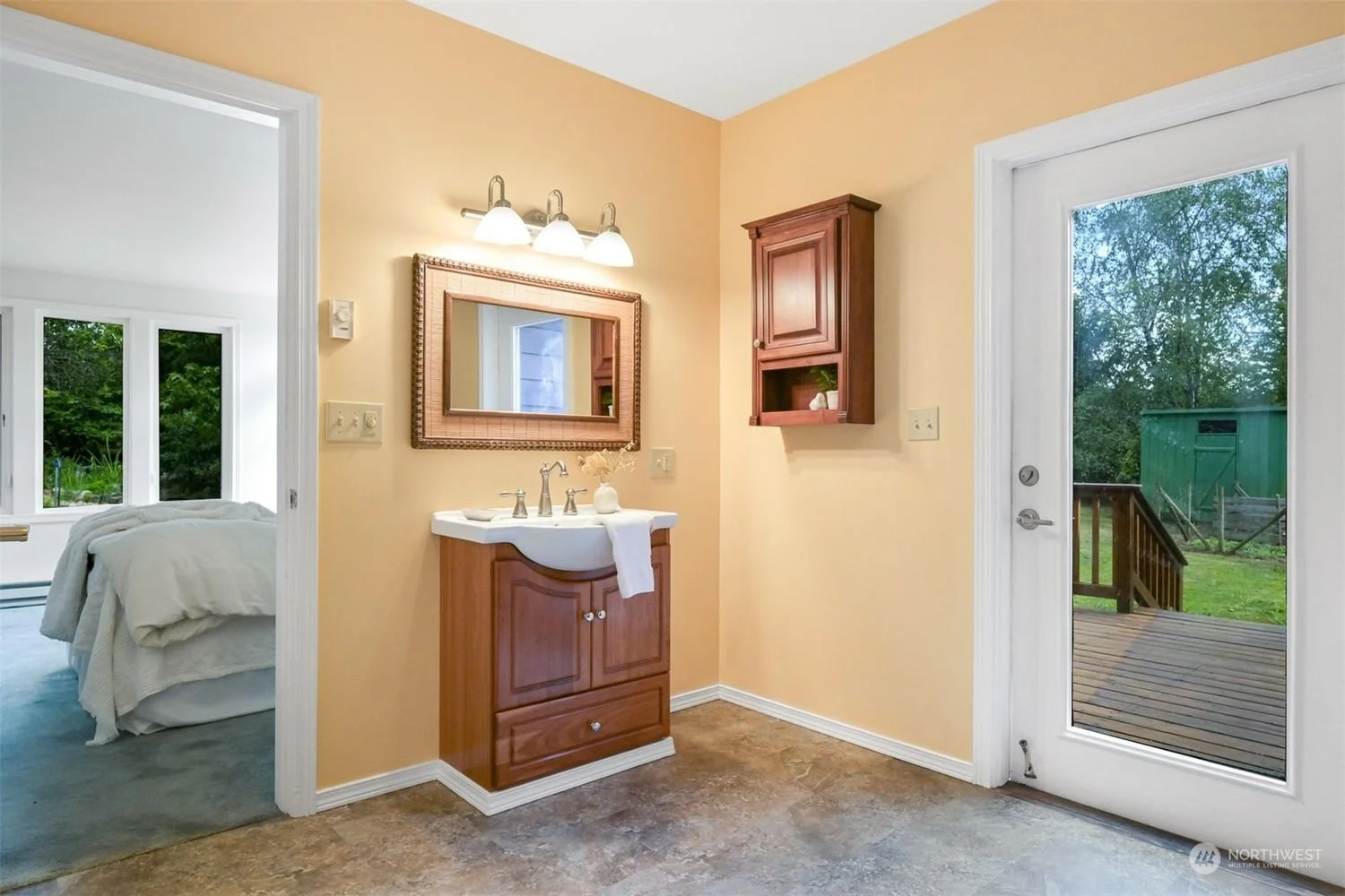 Bathroom with a small wooden vanity, mirror, wall-mounted cabinet, and a door leading to an outdoor deck with green yard and trees visible outside.