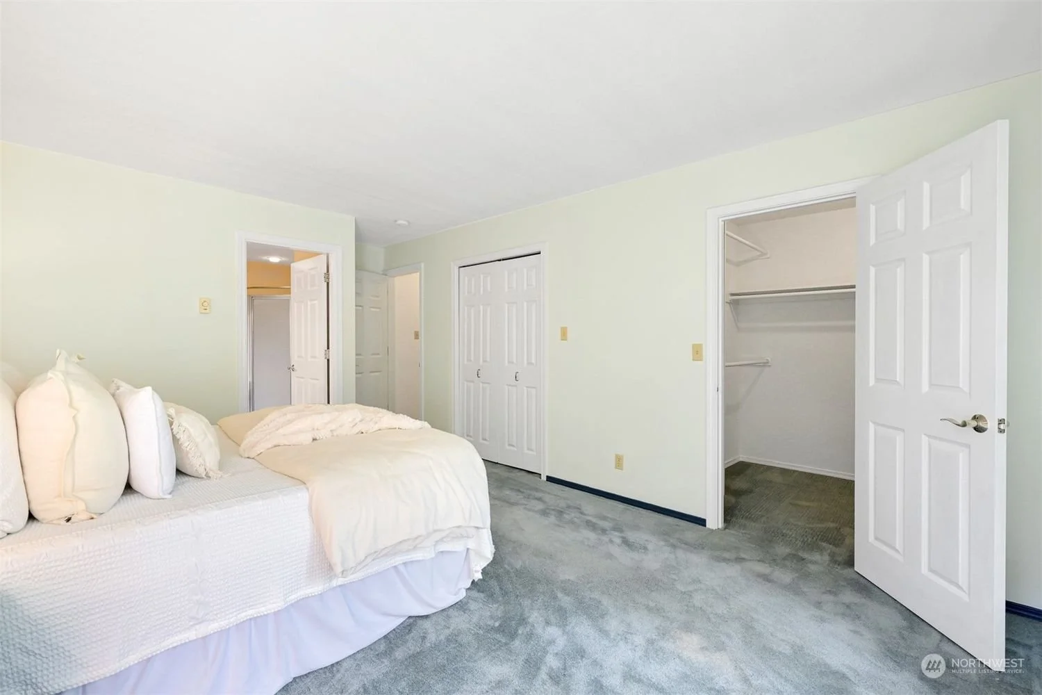 Empty bedroom with a bed on the left, open closet door on the right, and three doors in the background, all with white doors and light green walls.