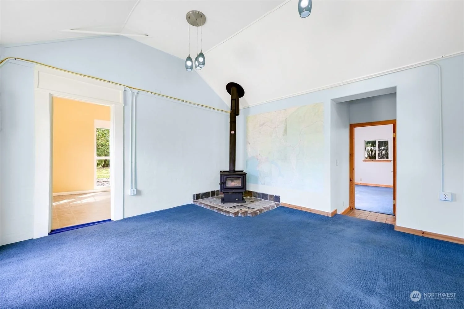 Empty living room with blue carpet, wood trim, a wood stove on a brick platform, and two doorways leading to other rooms, one with a bright yellow wall.