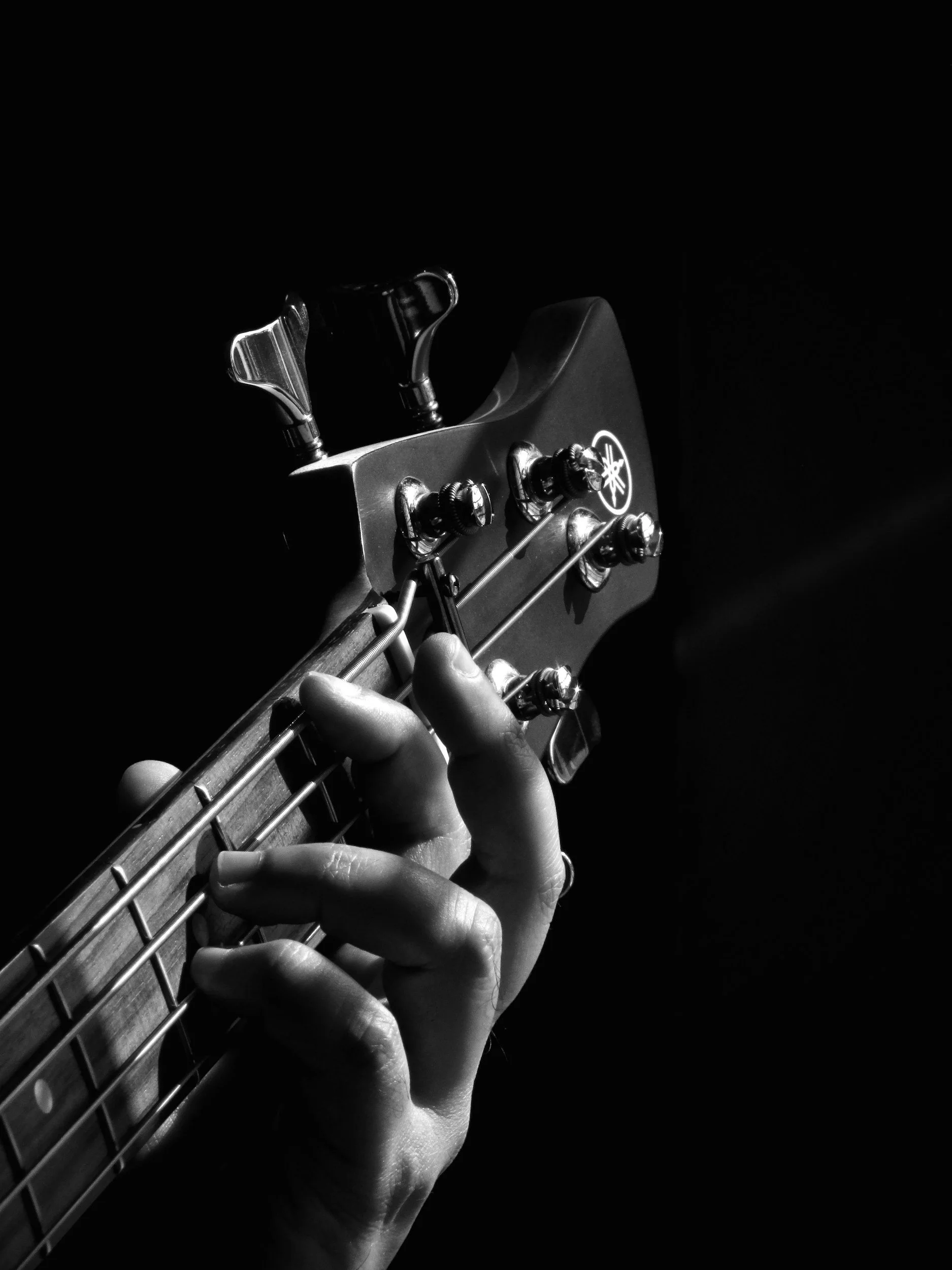 Close-up of a person's hand playing a Yamaha electric guitar. The image is in black and white, highlights the guitar's tuning pegs, headstock logo, and strings, with a dark background.