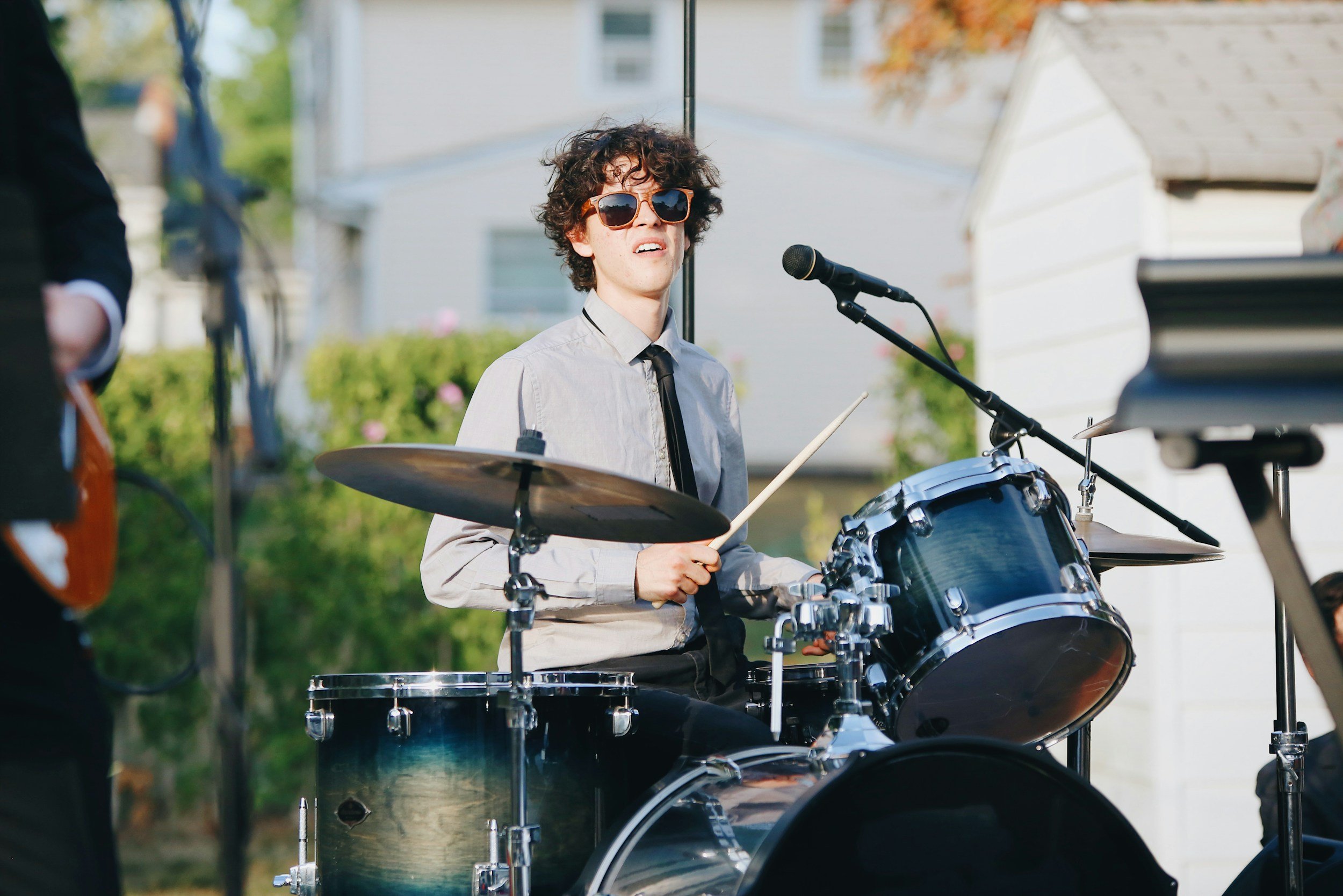 A young man with curly hair wearing sunglasses, a light-colored shirt, and a black tie, playing a drum set outdoors during daytime, with a microphone in front of him and other musicians partially visible.