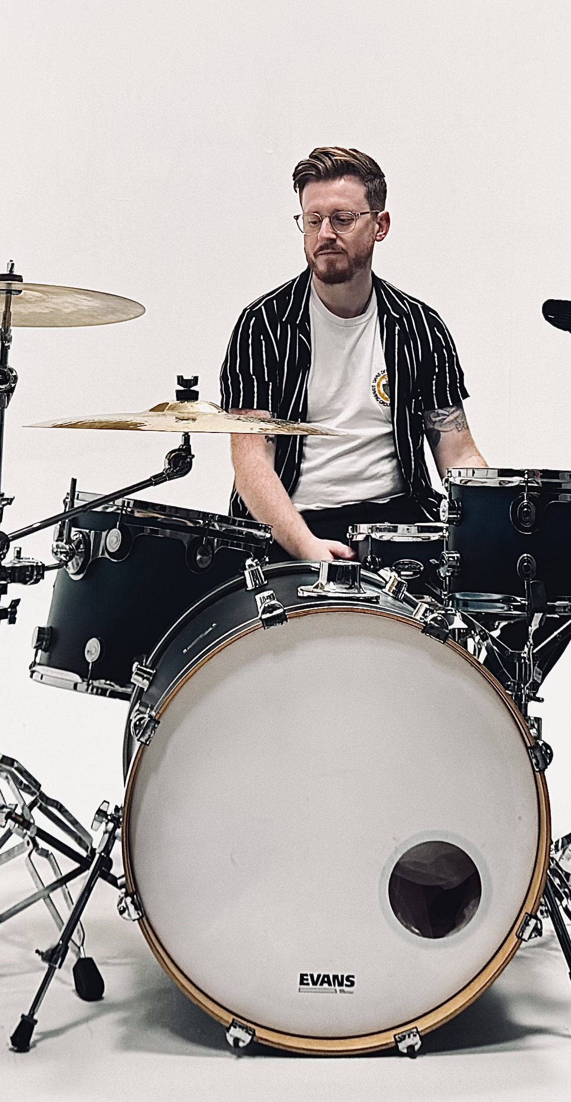 A man with glasses and tattoos sitting behind a black drum set with a white bass drum, against a plain white background.