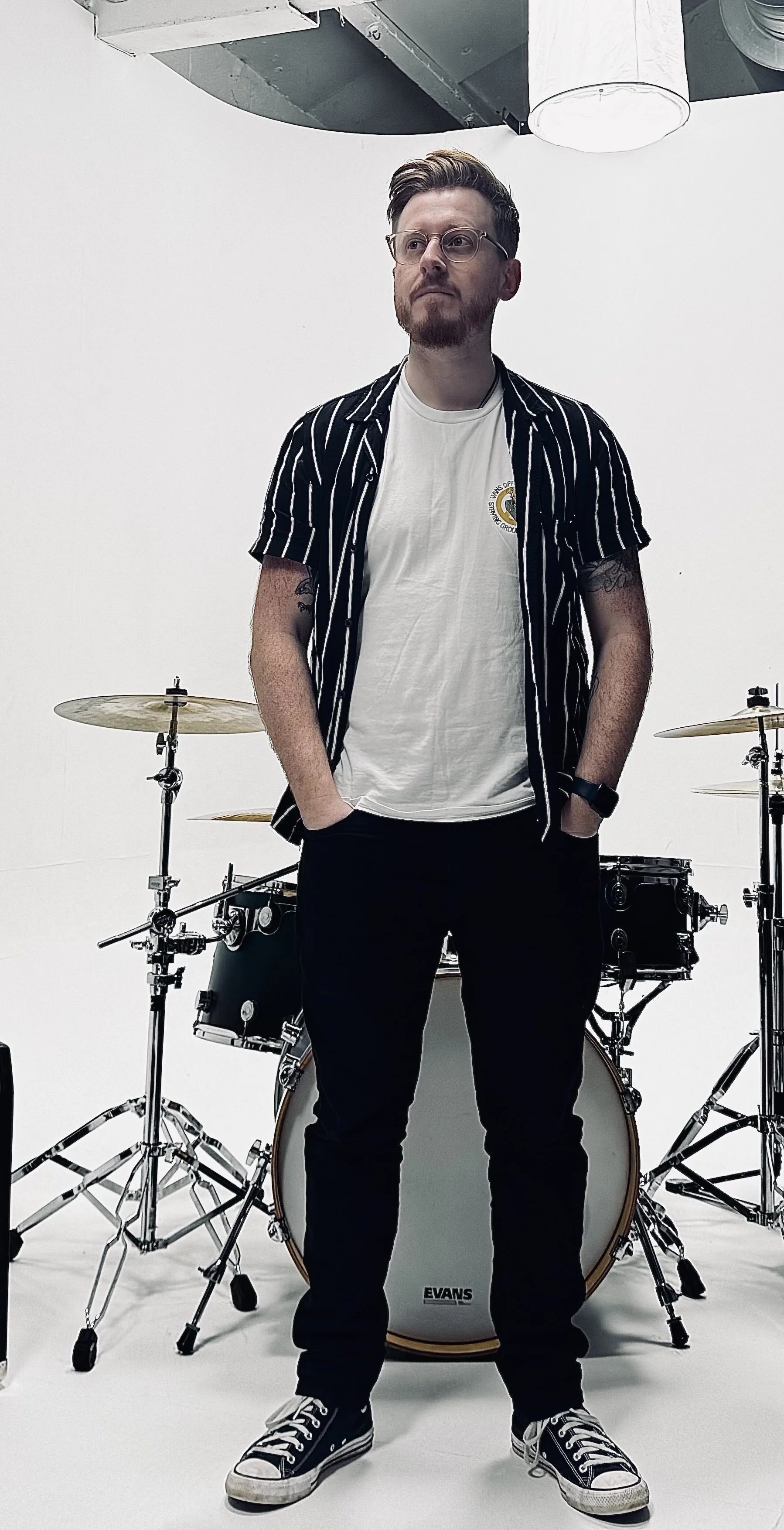 A young man with glasses and a beard stands with hands in pockets in front of a drum set, wearing a black and white striped shirt, white t-shirt, black pants, and sneakers, in a studio with a white background and ceiling lights.