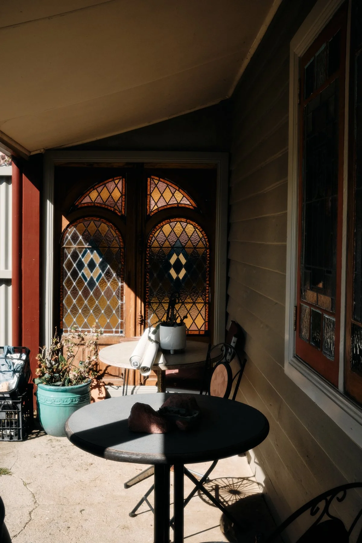 A shaded outdoor porch area with a round table, chairs, and a decorated window with stained glass and diamond patterns. There are potted plants, rolled-up papers, and a small crate on the porch.