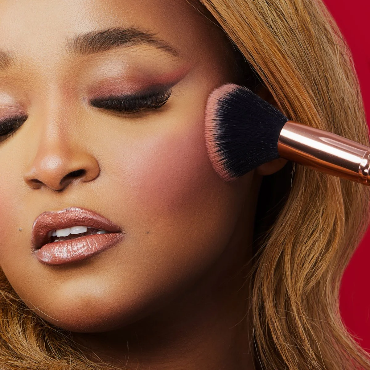 A woman with makeup applying blush using a large makeup brush on her cheek, with closed eyes and a red background.