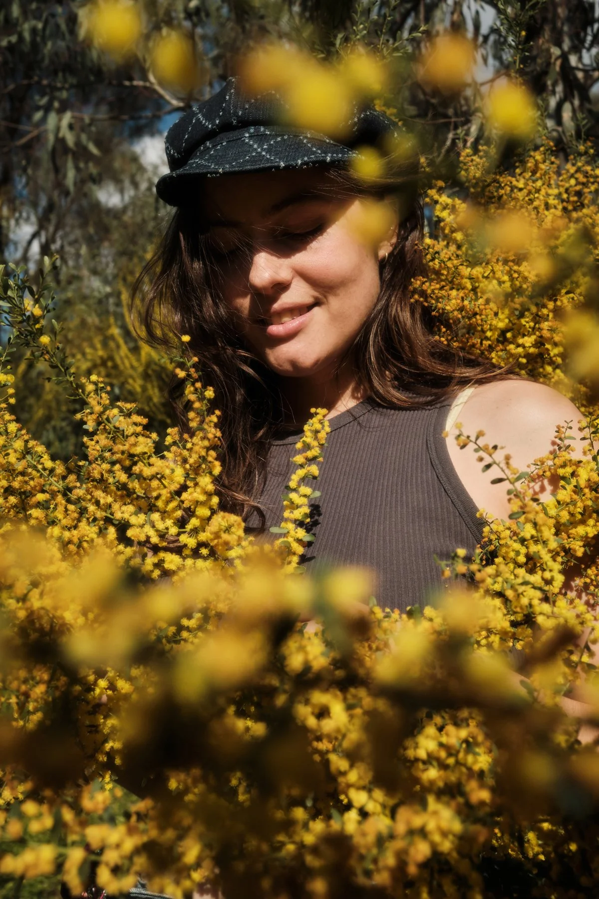 A woman with long dark hair, wearing a black cap and a sleeveless shirt, smiling gently among yellow flowering bushes outdoors.