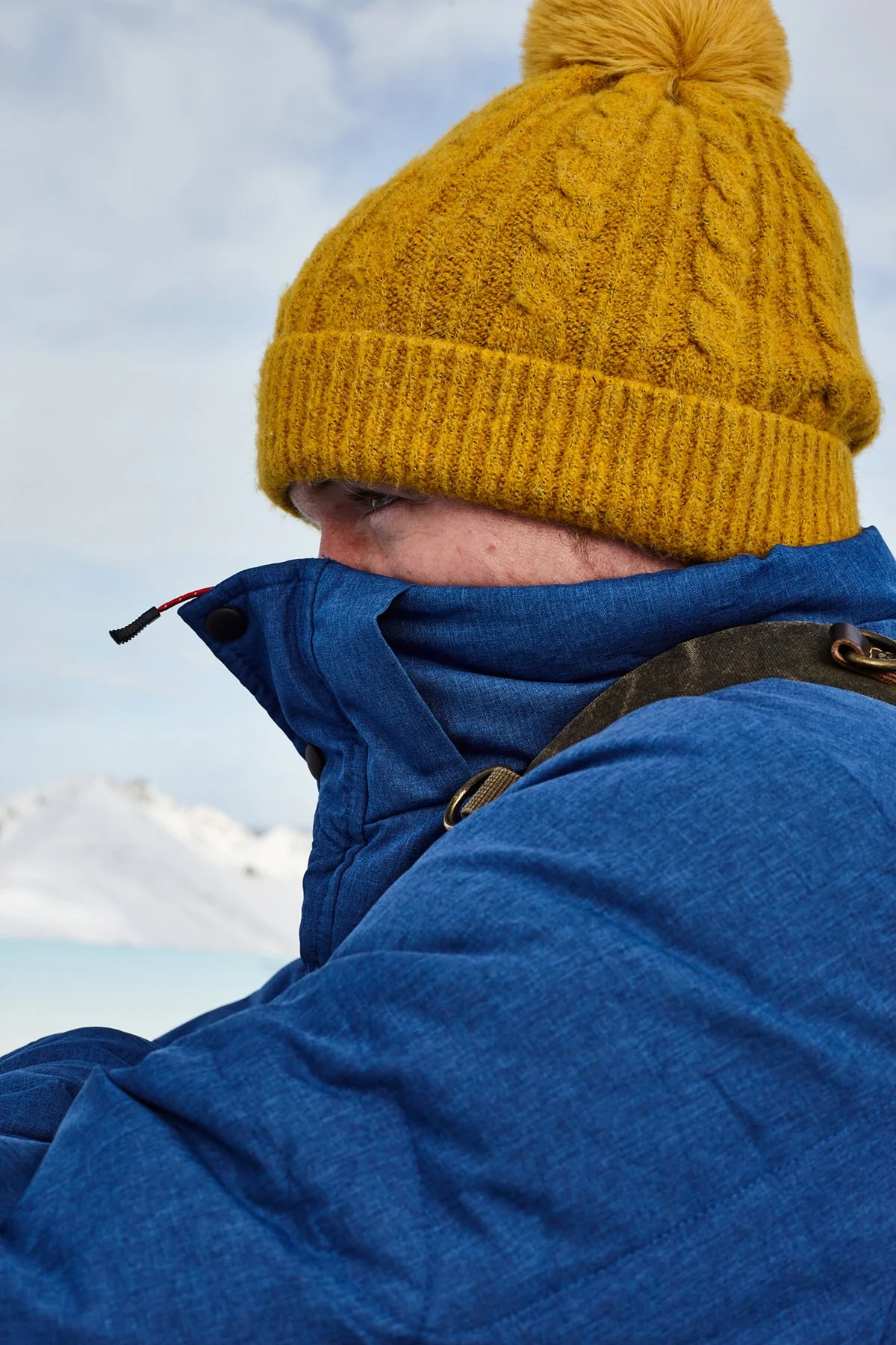A person bundled up in a blue outdoor jacket with the collar pulled up, wearing a mustard yellow knit beanie with a pom-pom on top, snow-covered mountains in the background.
