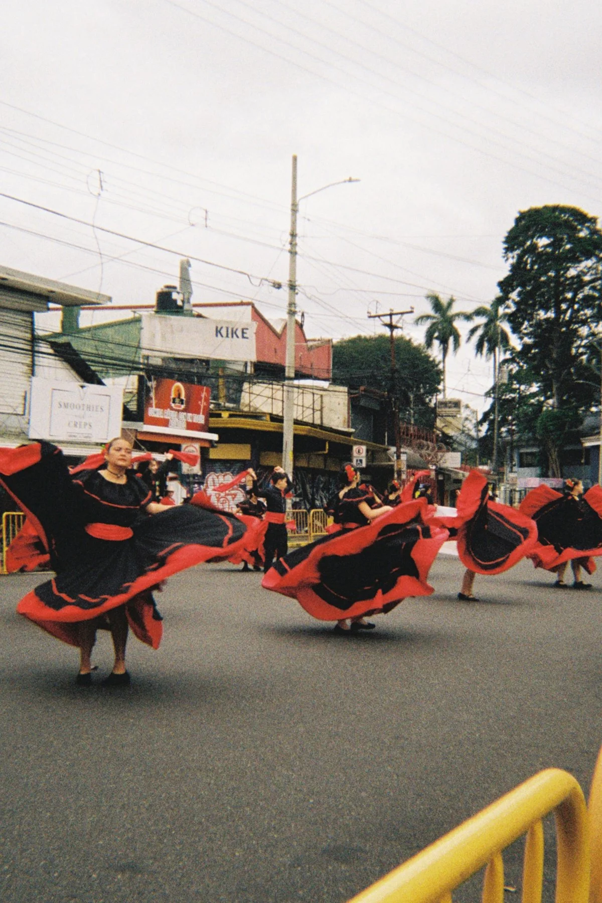 Group of women in black and red traditional dresses dancing in the street during a parade, with shops and trees in the background.