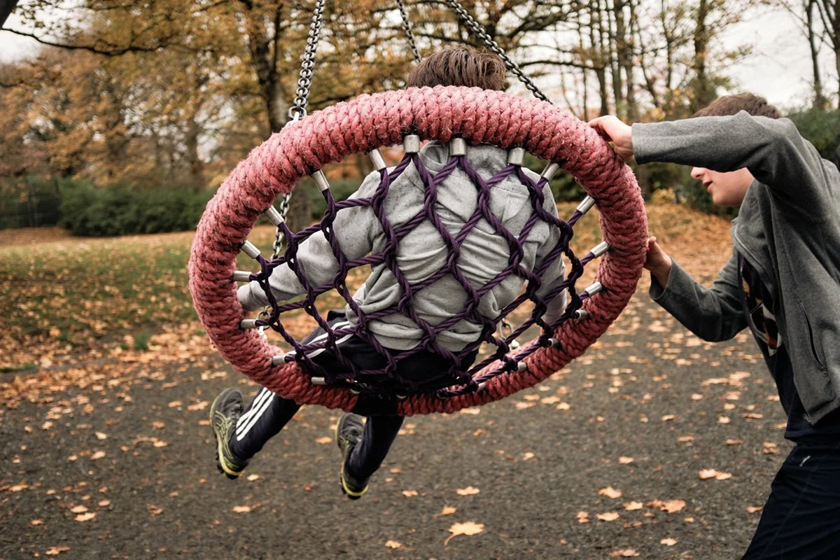 Children playing on a playground swing in fall with trees and fallen leaves in the background.