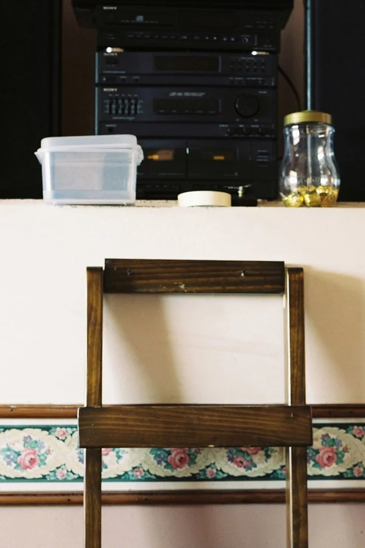 A wooden chair in front of a white cabinet with items on top, including a black stereo system, a plastic container, a jar with a golden lid filled with capsules, and a small round white object.