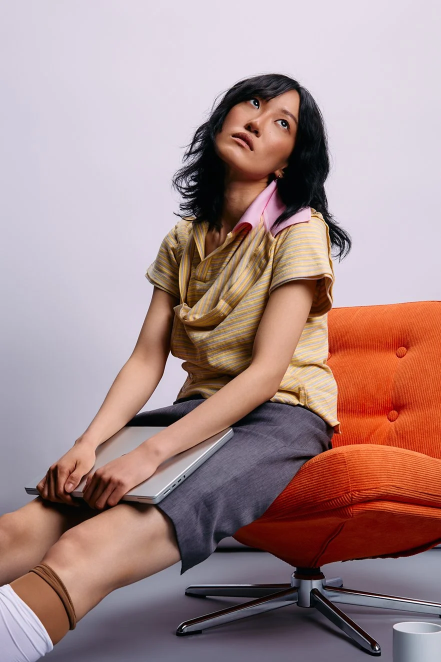 Fashion photography of a woman with black wavy hair wearing a yellow striped shirt and gray skirt sitting on an orange chair, holding a closed laptop on her lap, with a bored expression.