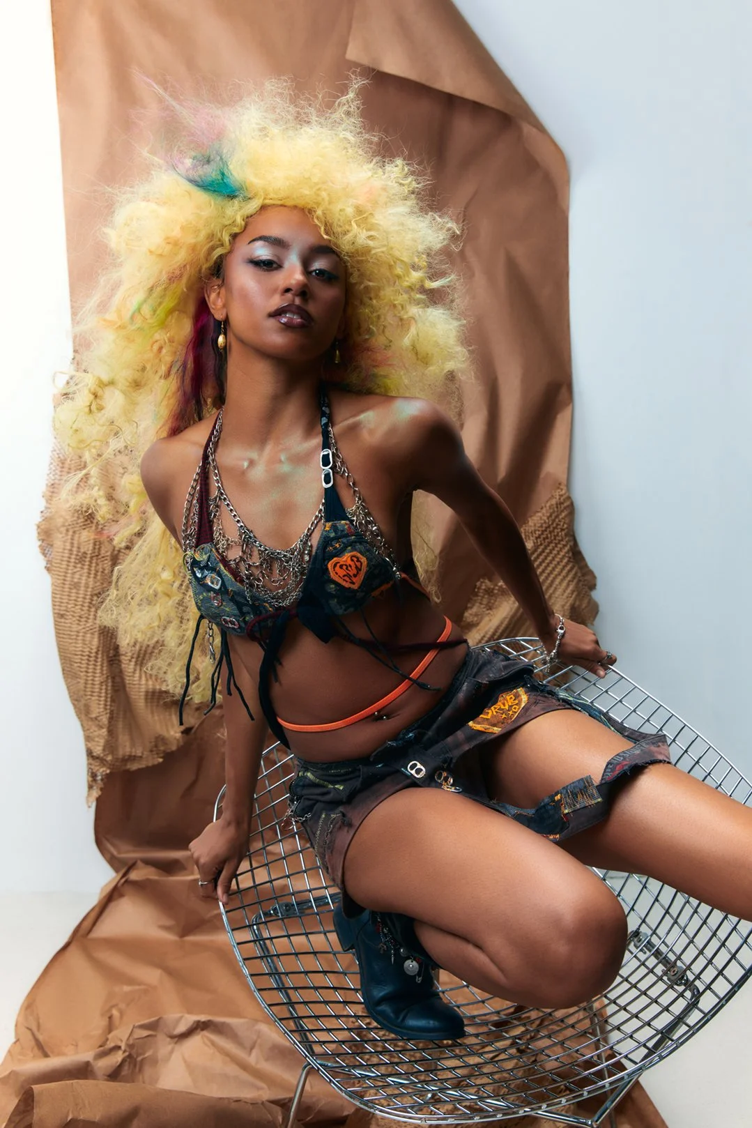 Fashion photograph of a woman with colorful, frizzy yellow hair with pink and blue streaks, wearing a distressed outfit, and black boots, sitting on a metal wire chair with a brown paper backdrop.