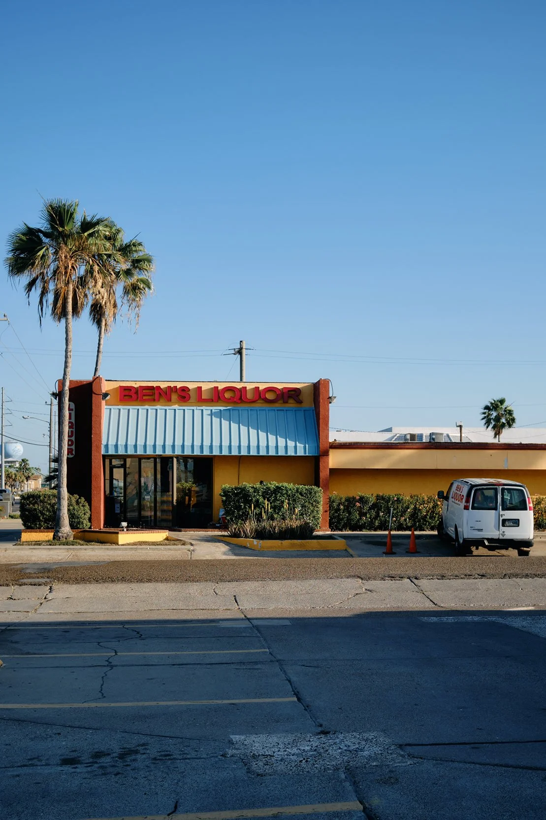 Travel photography of a liquor store named 'Ben's Liquor' with a yellow building, red sign, blue awning, palm trees, and a white van parked in front.