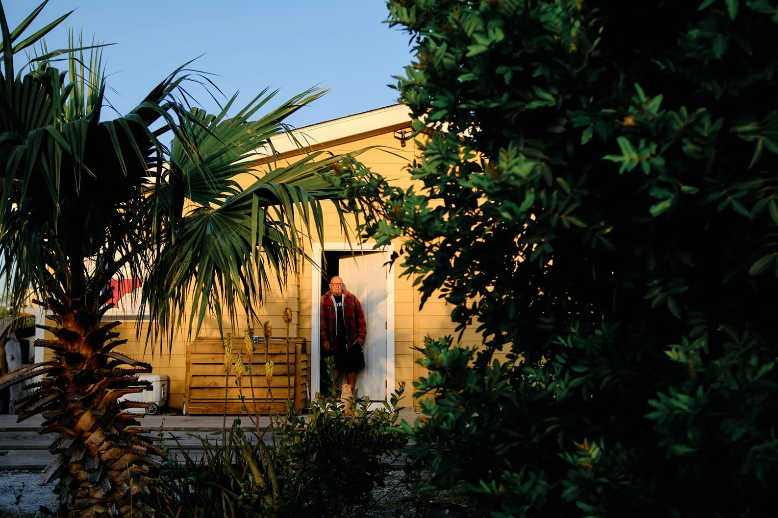 Travel photography of a man wearing a red plaid shirt, black shorts, and glasses standing in a doorway of a yellow house, surrounded by green tropical plants and trees, during the daytime.