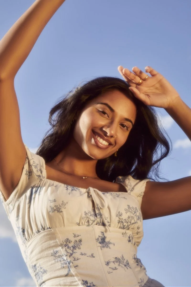 A woman smiling and posing outdoors against a clear blue sky, wearing a light-colored dress with a floral pattern.
