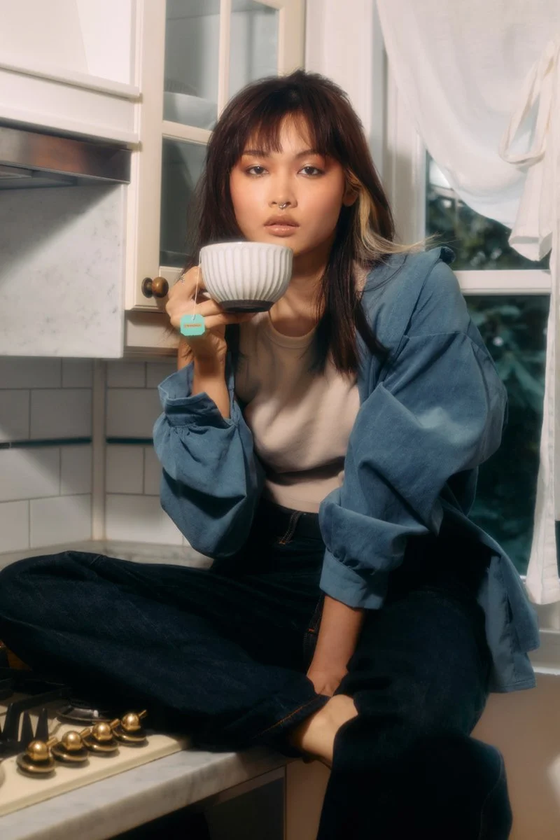 A young woman with dark hair, styled with blonde highlights, sitting cross-legged on a kitchen counter, holding a striped white bowl near her face, wearing a beige shirt and a blue denim jacket, with a window and kitchen cabinets in the background.