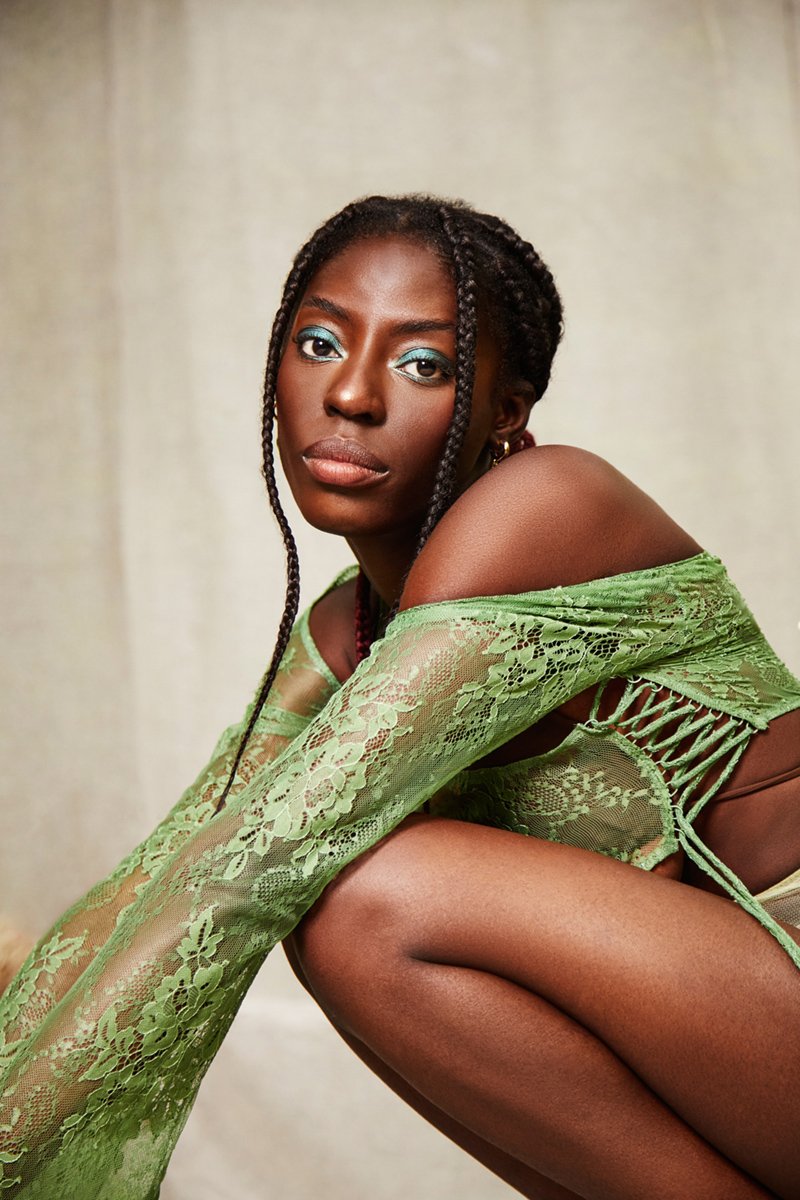 A woman with braided hair and bold makeup is sitting with one knee up, wearing a green lace top that exposes her shoulder. The background is plain and light-colored.