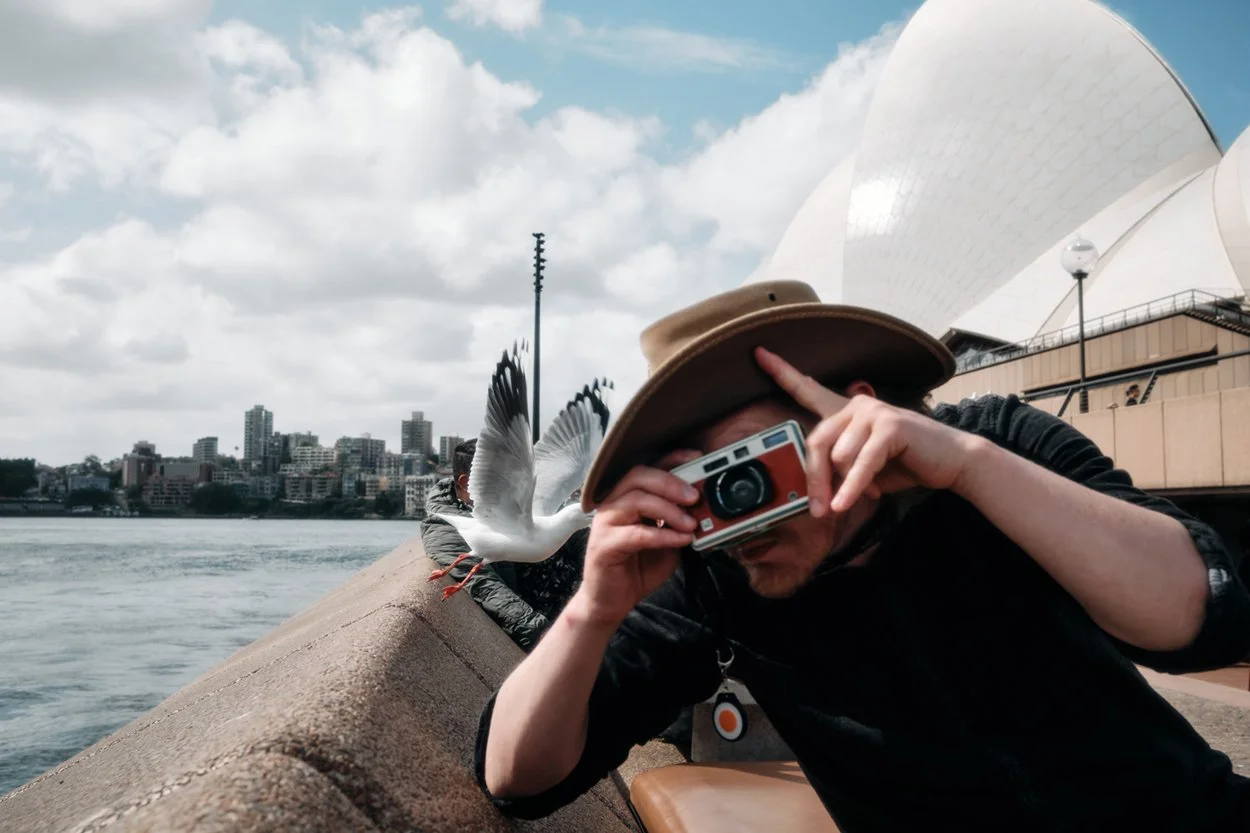 A person wearing a brown hat and black jacket taking a picture with a vintage camera, with a seagull flying nearby, on a riverside promenade with the Sydney Opera House and city skyline in the background.