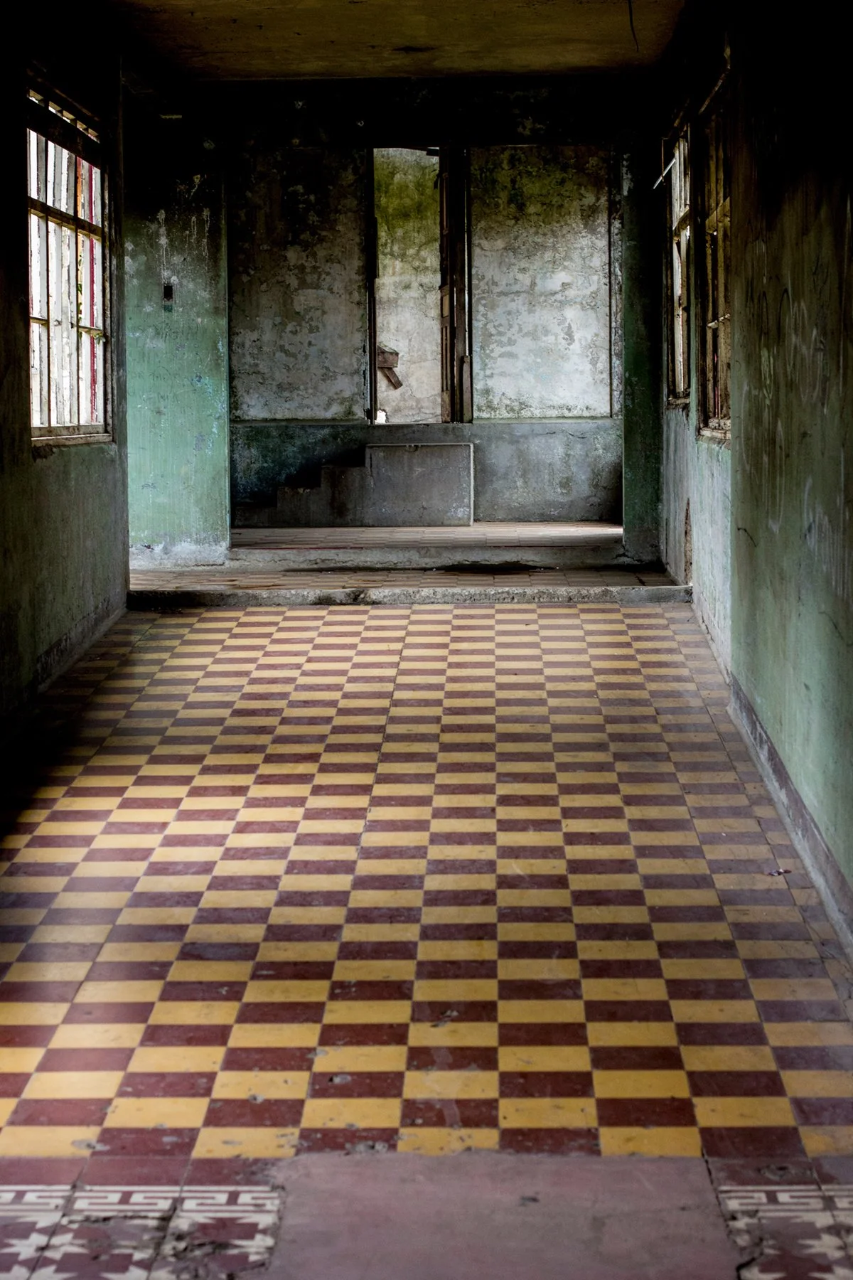 Interior of an abandoned building with peeling paint, broken windows, and a checkered yellow and red tile floor.