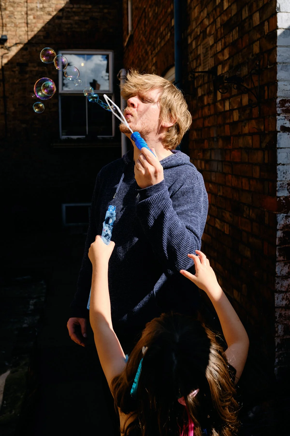 A man blowing bubbles while a young girl reaches up to touch them outside near brick buildings.