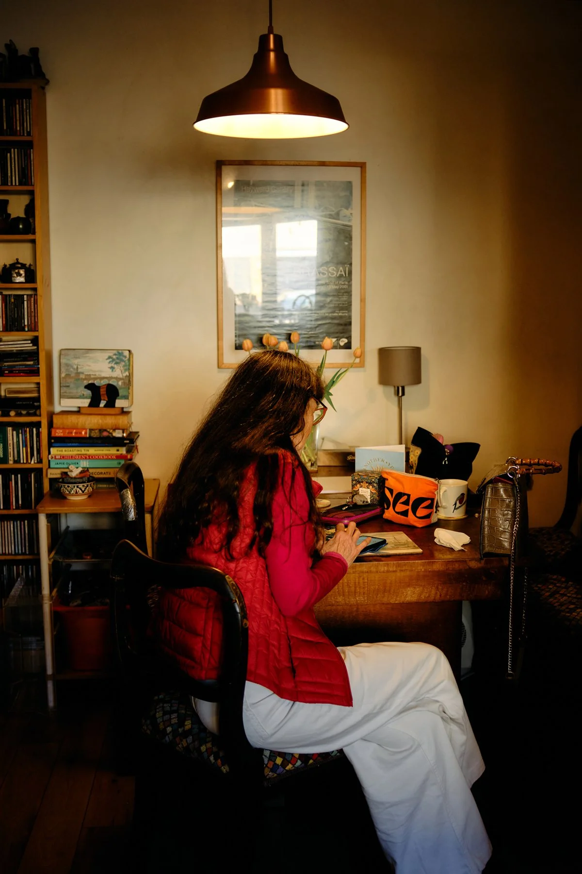 A woman with long dark hair and glasses sitting at a wooden table, using a smartphone, in a cozy room with a bookshelf, framed posters, and a lamp overhead.