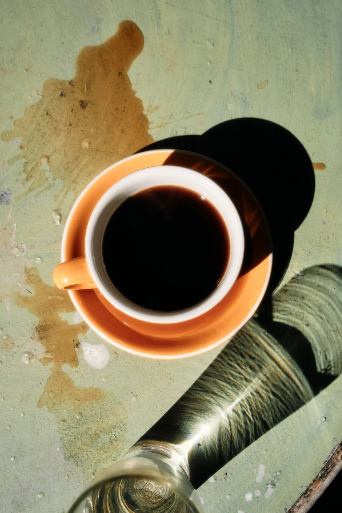 A cup of black coffee in an orange cup and saucer, seen from above, with a glass bottle and coffee spills on a green surface.