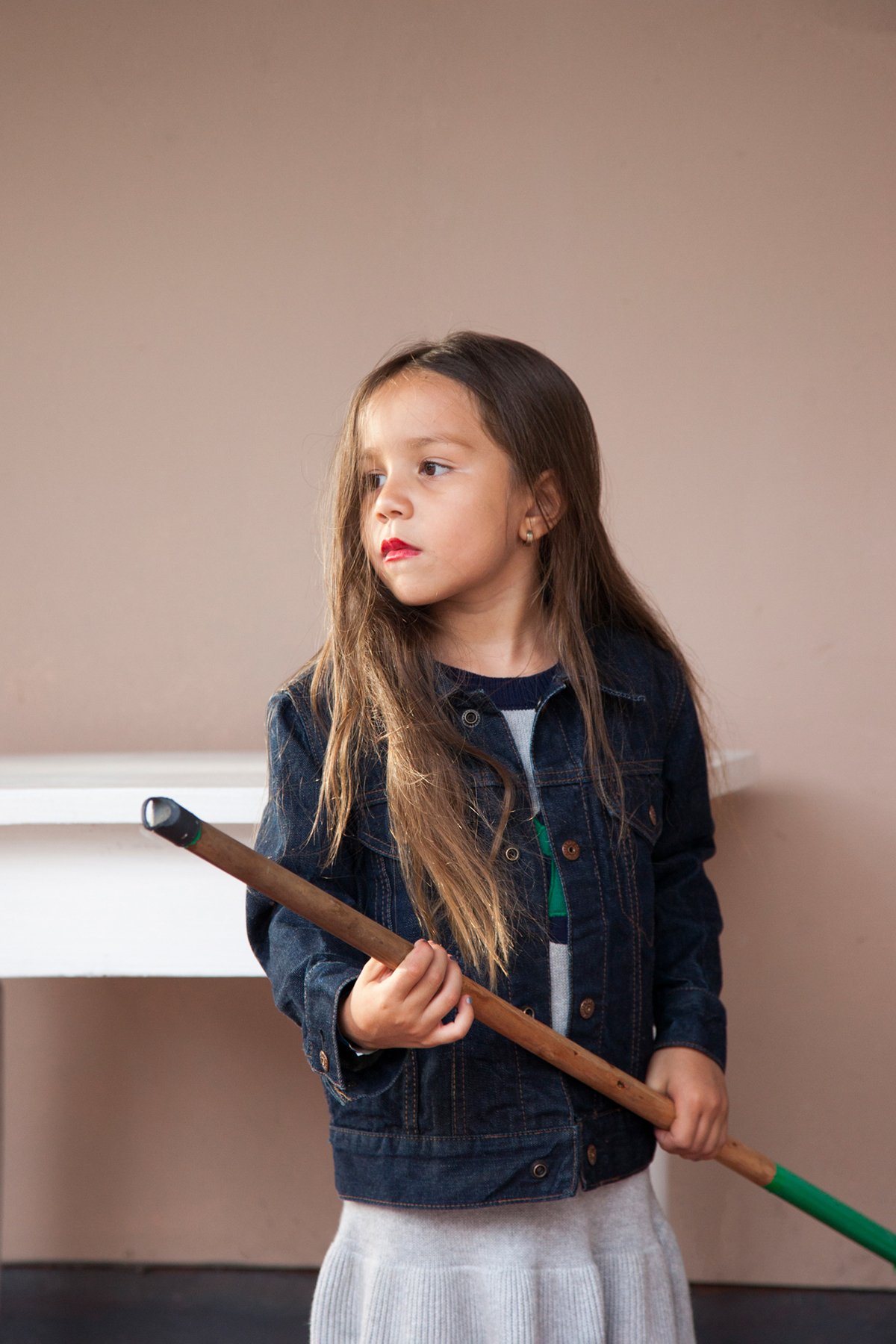 A young girl with long brown hair holding a pool cue stick, standing in a room with a plain beige wall and a white table in the background.
