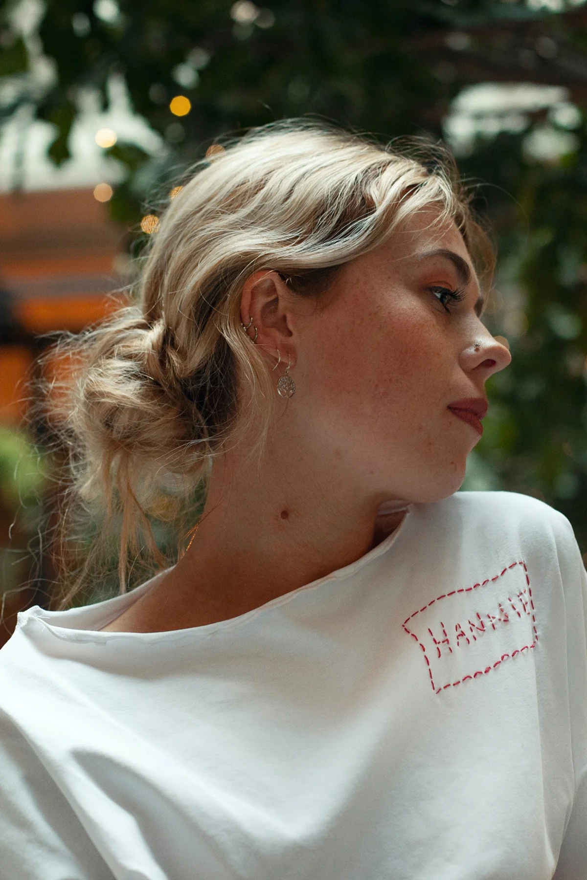 Profile of a young woman with blonde hair styled in a loose bun, wearing earrings and a white top with red stitching that reads 'THANK'.