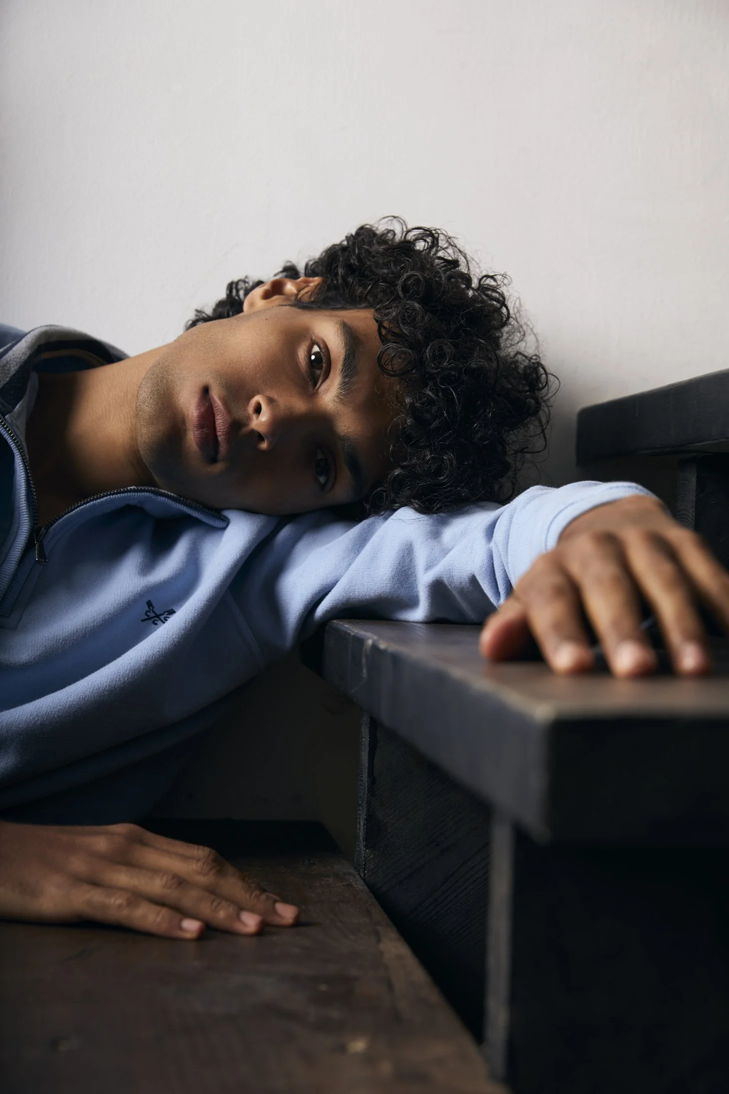 A young man with curly black hair and brown skin laying on a wooden table, resting his head on his arm, with a plain white wall in the background for portrait photograph.