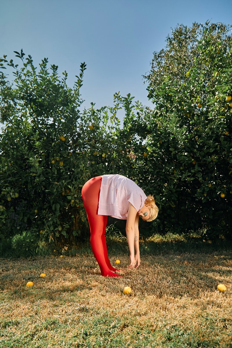 A woman bending over in an orchard with lemon trees, picking up a yellow lemon, while several lemons are on the ground.