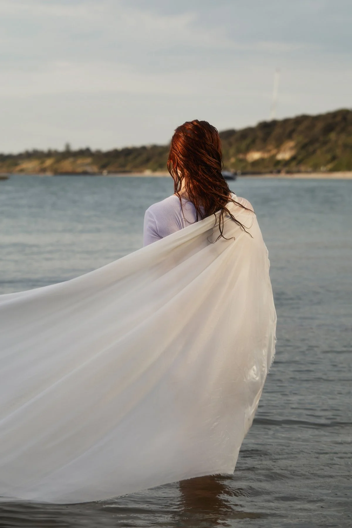 A woman with long, wet red hair, standing in the water near a shoreline, wearing a white dress or gown with a long train flowing behind her, facing away from the camera.