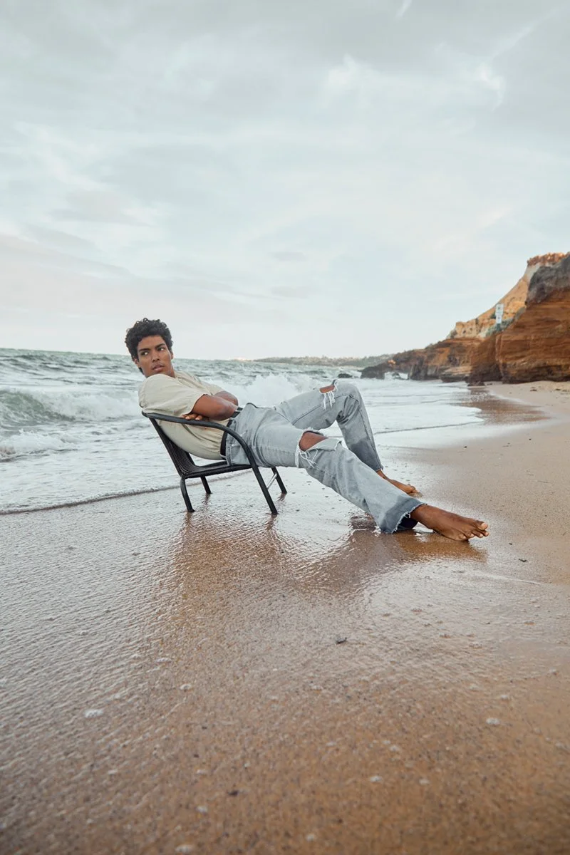Young man sitting on a chair at the beach with waves crashing nearby and rocky cliffs in the background.