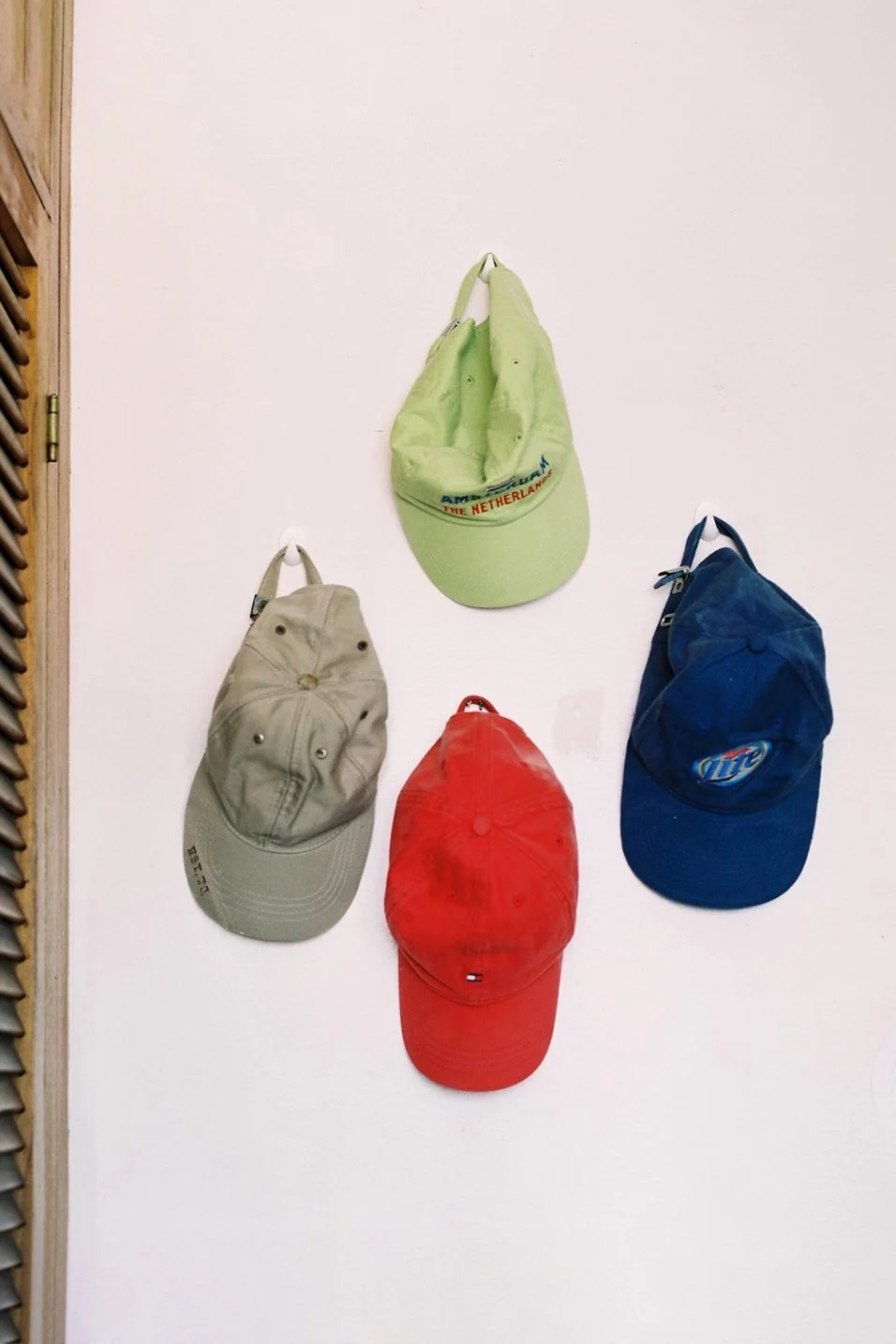 Four baseball caps hanging on a white wall, each in different colors: beige, red, green, and blue.