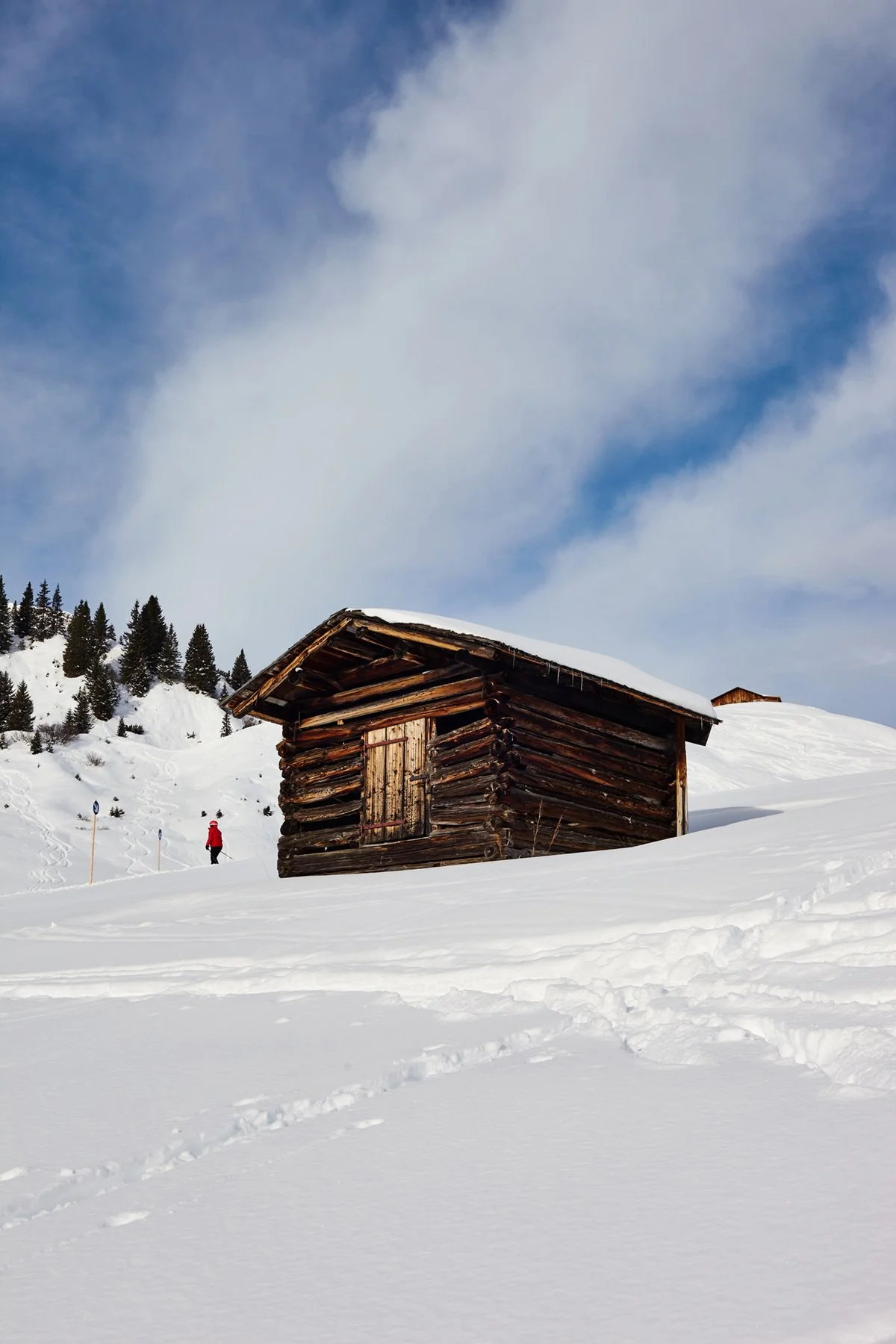 A snowy landscape with a rustic wooden cabin and a person in a red jacket walking in the background, surrounded by snow-covered trees under a partly cloudy sky.