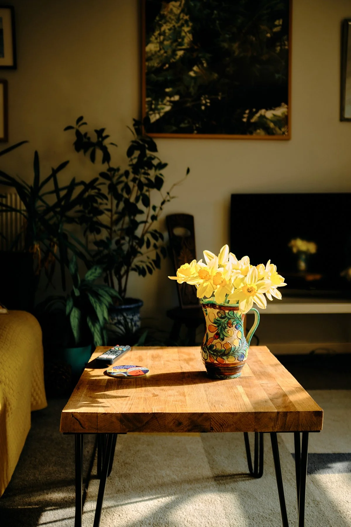 A wooden coffee table with a colorful ceramic vase filled with yellow daffodils, located in a cozy living room with a yellow sofa, potted plants, wall art, a television, and a remote control.