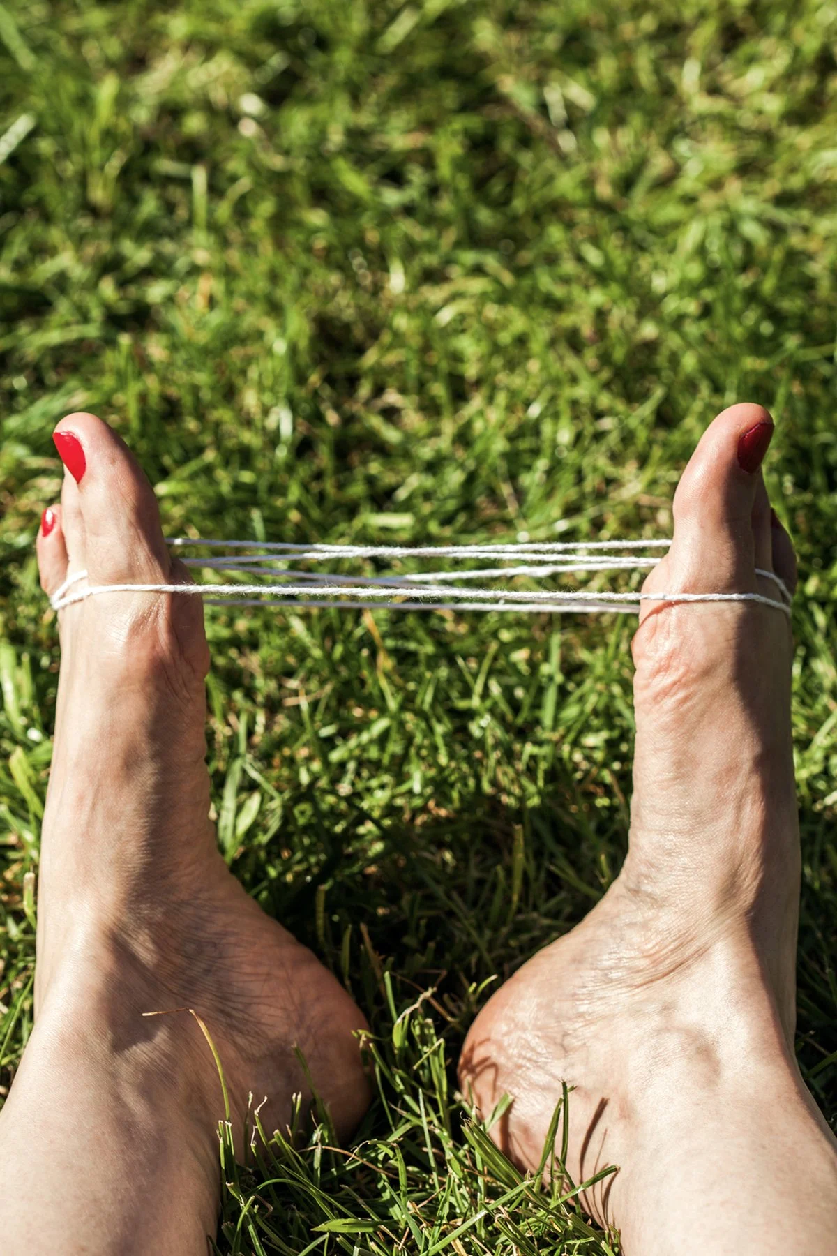 Person's feet with red toenail polish holding three loose white strings tied around the toes, with green grass in the background.