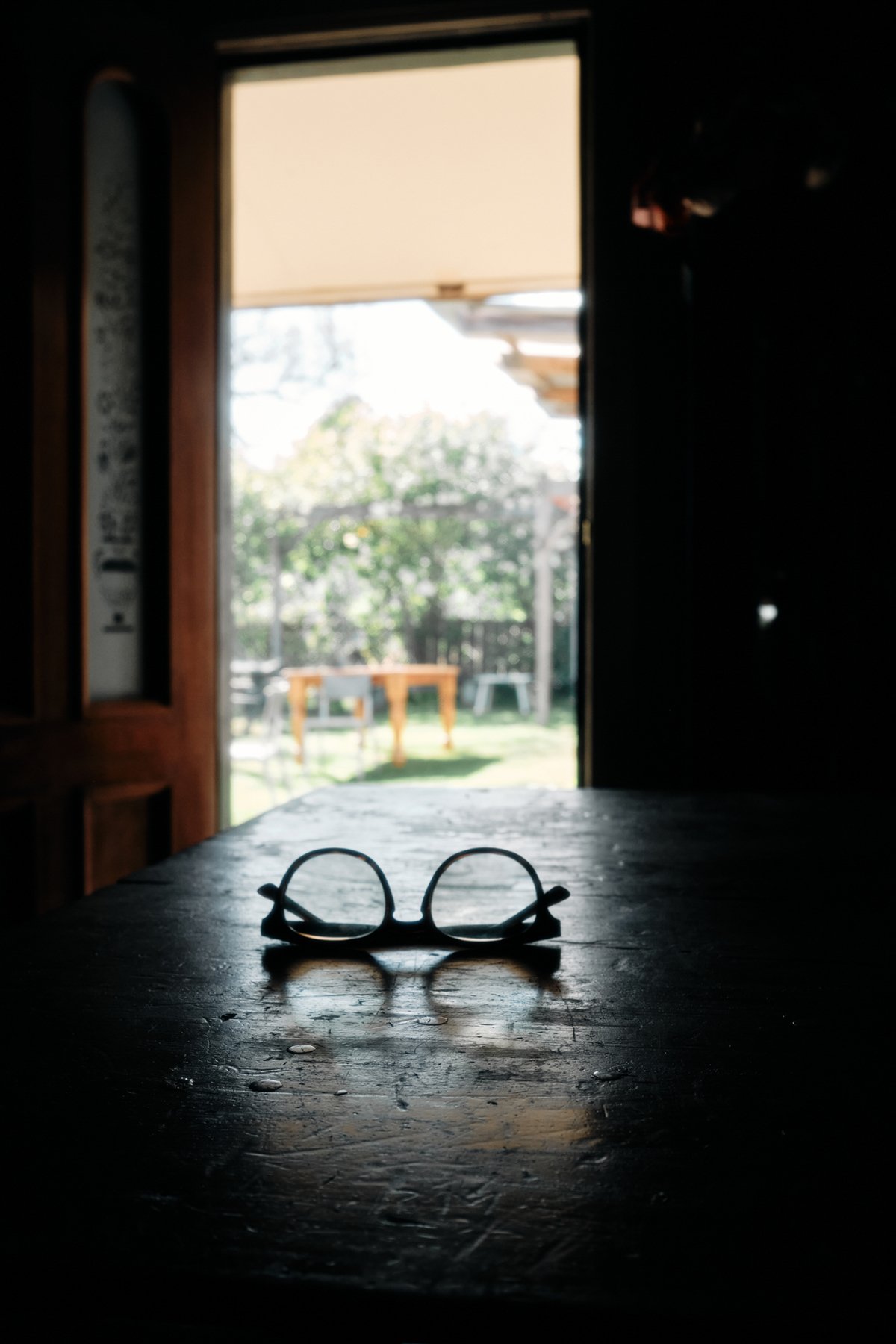 A pair of glasses on a dark wooden table inside a room, with sunlight coming through an open door leading outside to a garden with trees and a table.