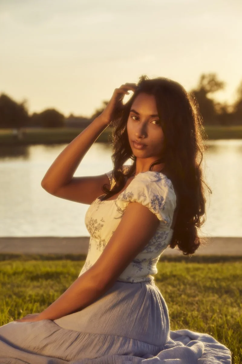 A woman sitting on a blanket by a lake during sunset, with her hand in her hair and looking at the camera.