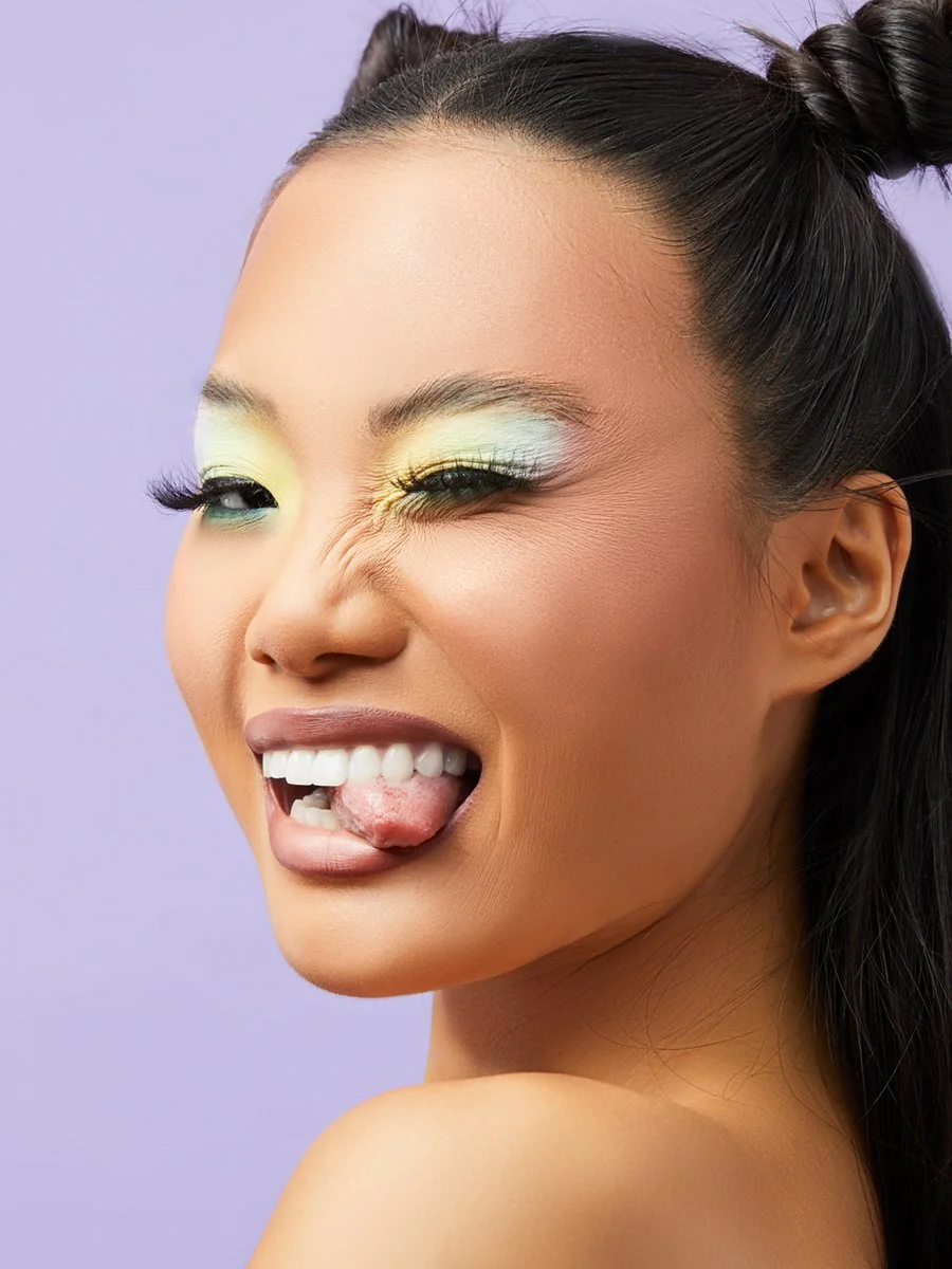A woman with colorful eye makeup sticking out her tongue and winking, against a purple background.