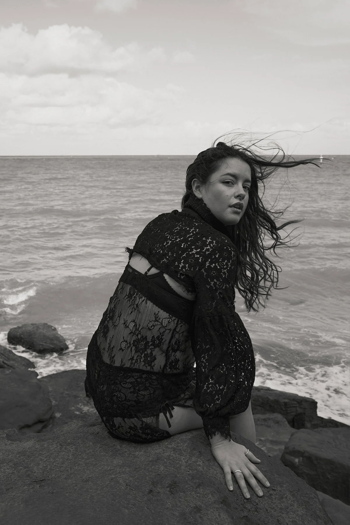 A woman with wavy hair sitting on rocks near the ocean, wearing a black lace dress, with her hair blowing in the wind, photographed in black and white.