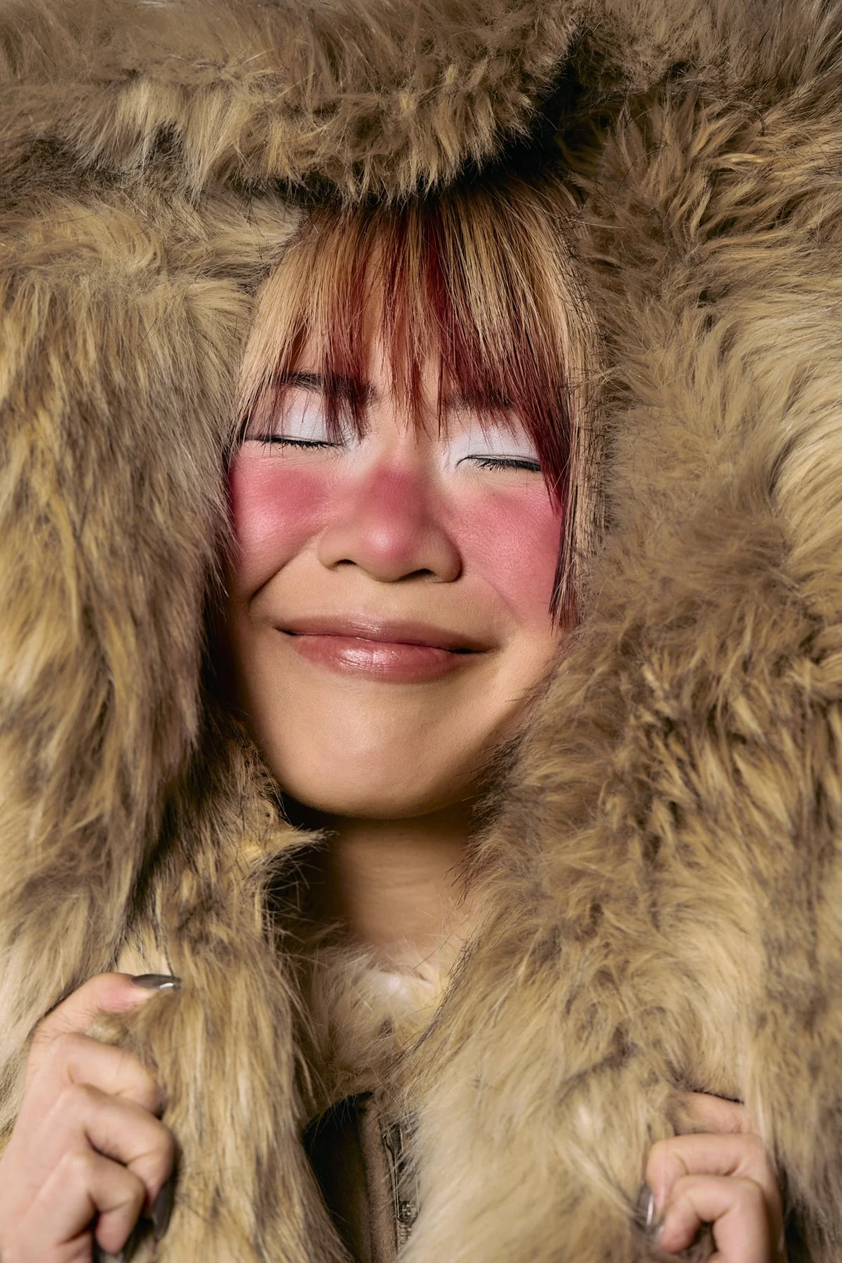 A woman with closed eyes, smiling, surrounded by a large fur coat or blanket.
