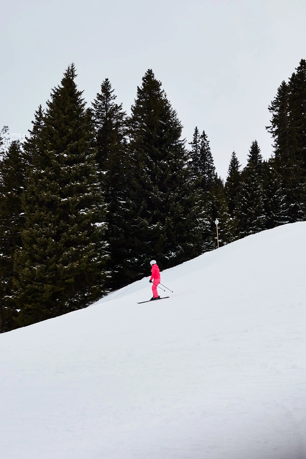 A person skiing down a snowy slope in winter, wearing pink clothing and a helmet, with pine trees in the background.