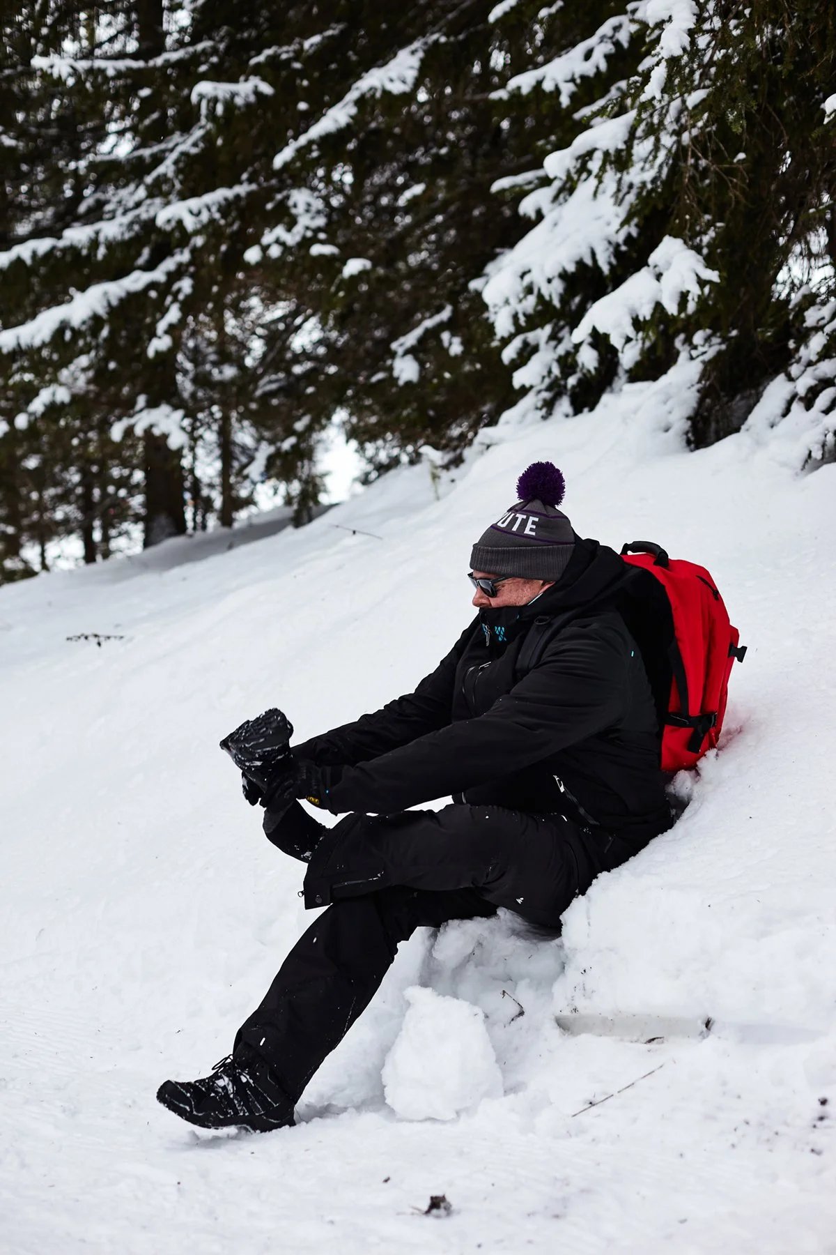 A man in winter clothing and sunglasses sitting on a snowbank in a snowy forest, adjusting his glove.